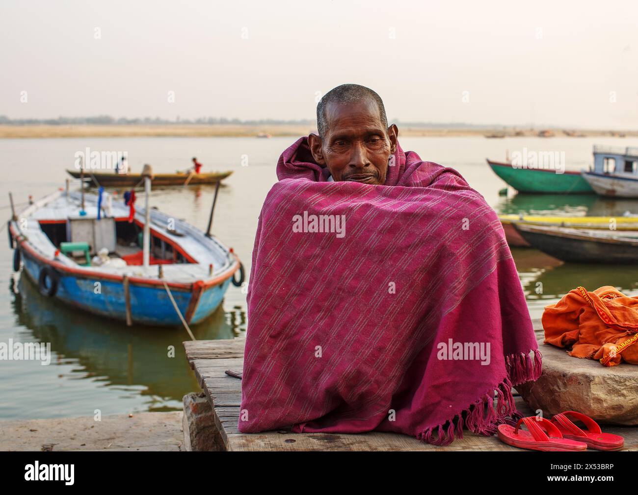 Mann, der mit Armen auf hochgezogenen Knien sitzt, umwickelt von einer kastanienbraunen Decke neben dem Ganges bei Varanasi, Indien. Stockfoto