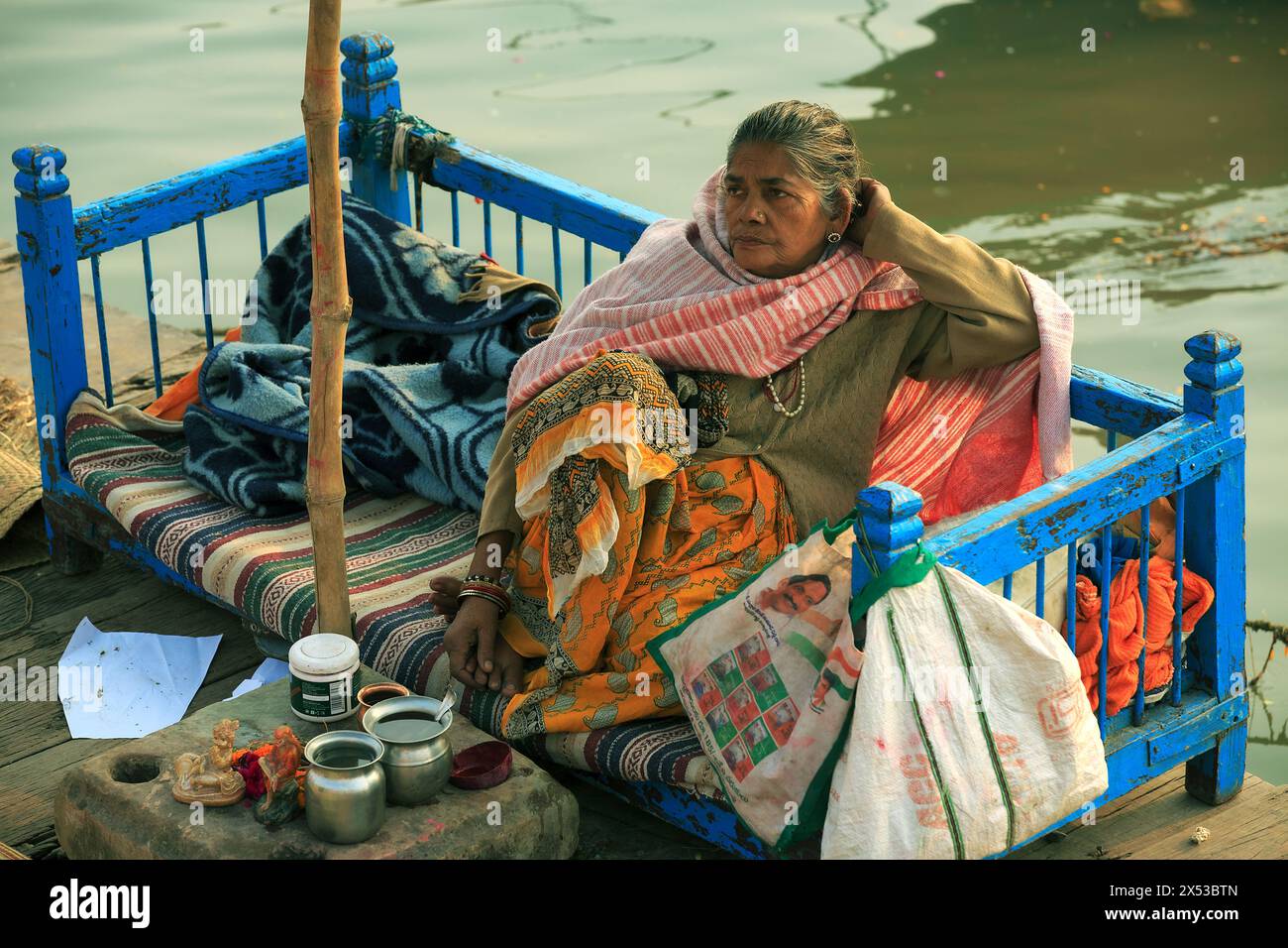 Hindu-Frau sitzt auf einer blau bemalten Holzbank mit Puja-Gegenständen vor ihr neben dem Ganges in Varanasi, Indien. Stockfoto