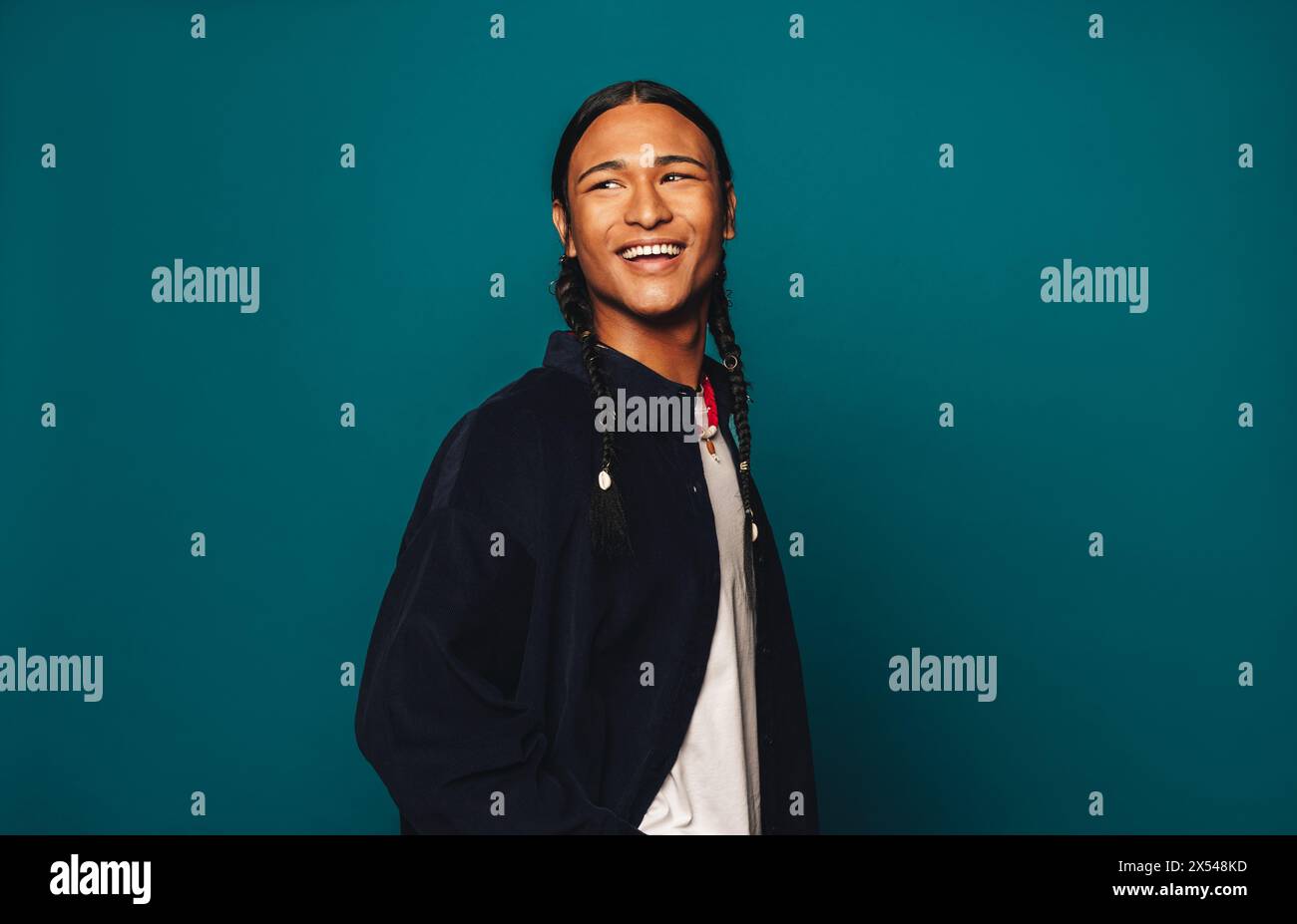 Junger Mann mit geflochtenen Haaren und einem glücklichen Lächeln steht im Studio mit einem lässigen und sorglosen Look. Geflochtenes Haar, das über seine Schultern kaskadiert, taucht er auf Stockfoto