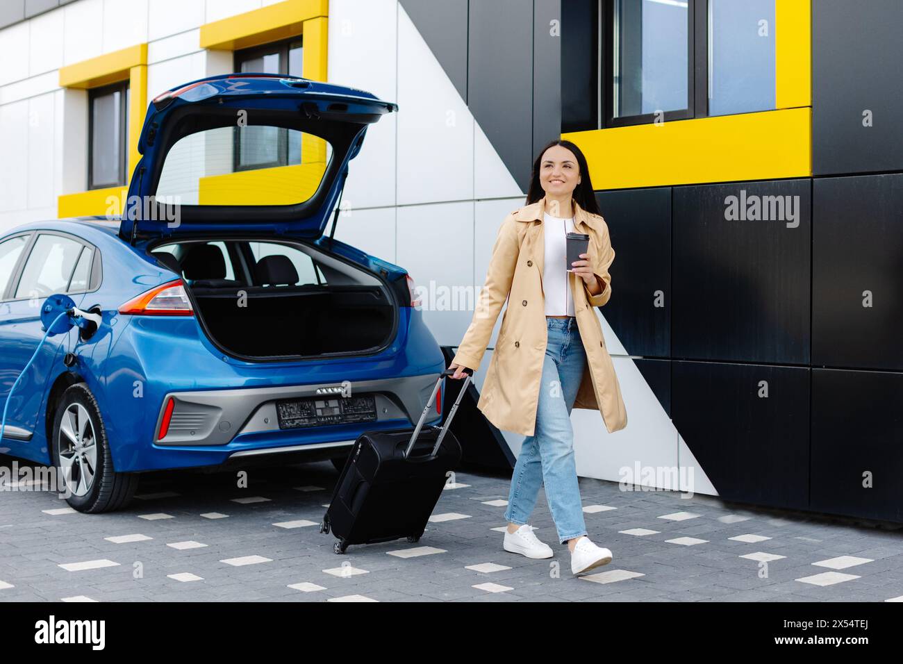 Ein junges Mädchen hielt an, um ihr Elektroauto aufzuladen, und in der Zwischenzeit ruht es sich aus und trägt einen Koffer. Stockfoto