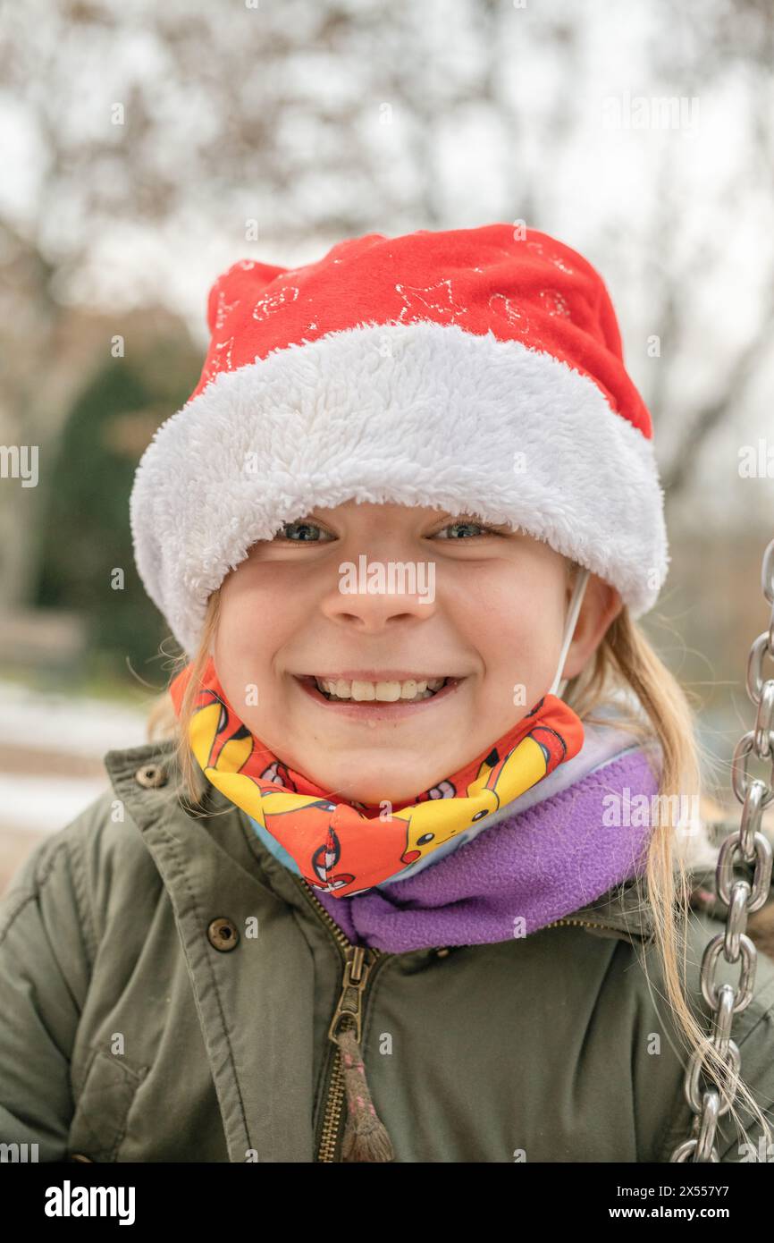 Freundliches, hübsches Mädchen auf einem Spielplatz mit einem schicken roten Weihnachtsmann-Hut. Im Winter, kurz vor Weihnachten, gibt es große Begeisterung über die Geschenke. Stockfoto