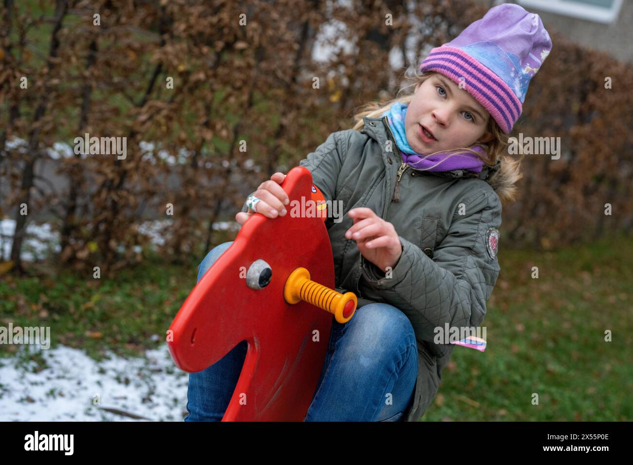 Mädchen lieben Pferde. Wenn kein Tier in der Nähe ist, spielen Kinder gerne auf einem Spielplatz mit einem roten Schaukelpferd. Gesund bei kaltem Wetter. Stockfoto