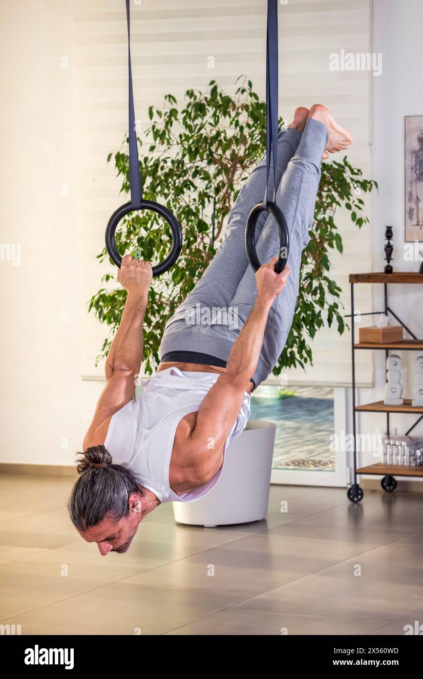 Seitenansicht eines starken erwachsenen Mannes mit Haargummi, der nach unten blickt, während er Calisthenics-Kernstabilitätsübungen an aufgehängten Ringseilen in Halle mit Daylig durchführt Stockfoto