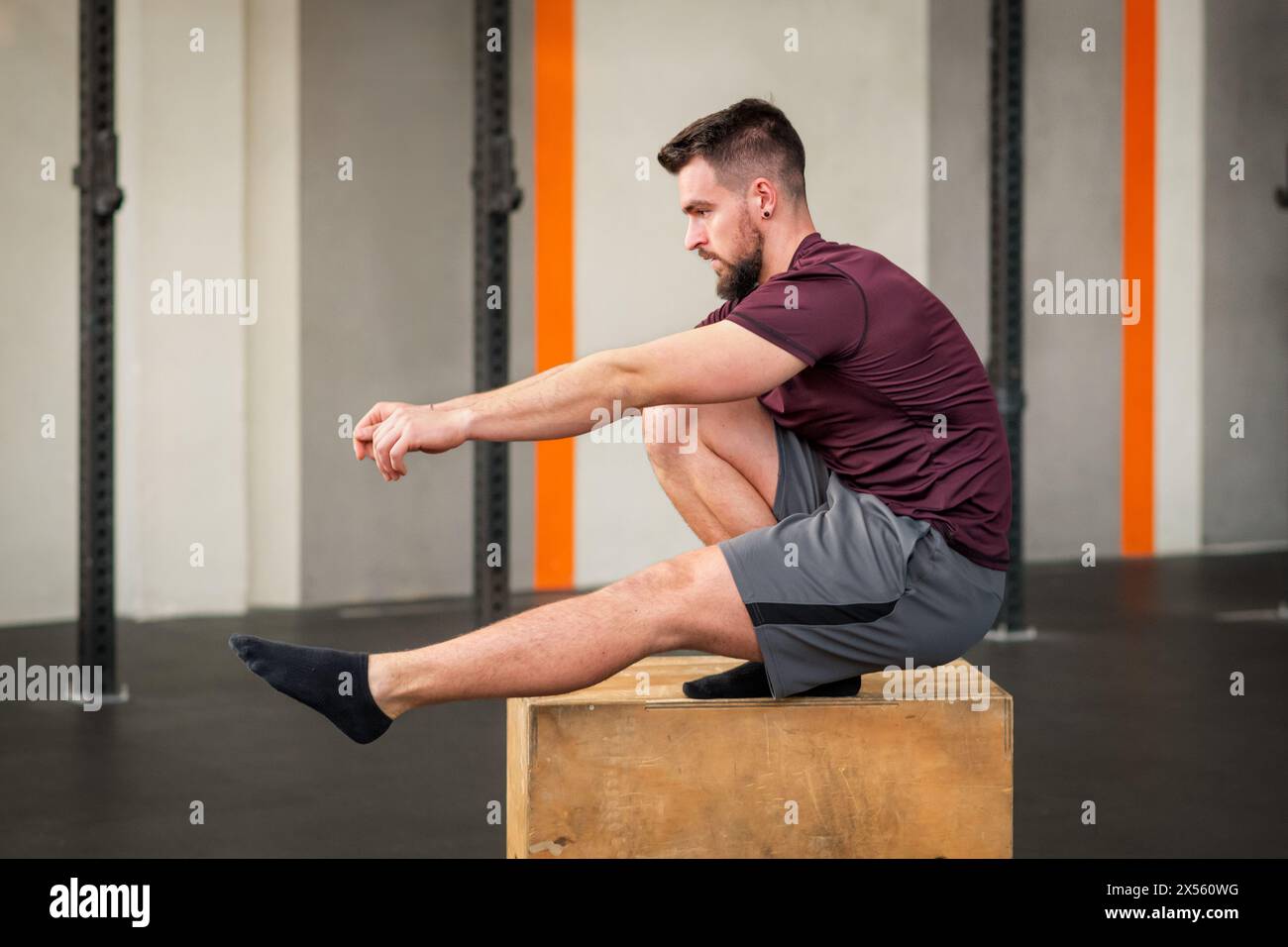 Seitenansicht eines starken bärtigen erwachsenen männlichen Athleten in Sportbekleidung, der wegschaut, während er auf der Box sitzt und Calisthenics-Pistolenhockübung bei Tageslicht macht Stockfoto