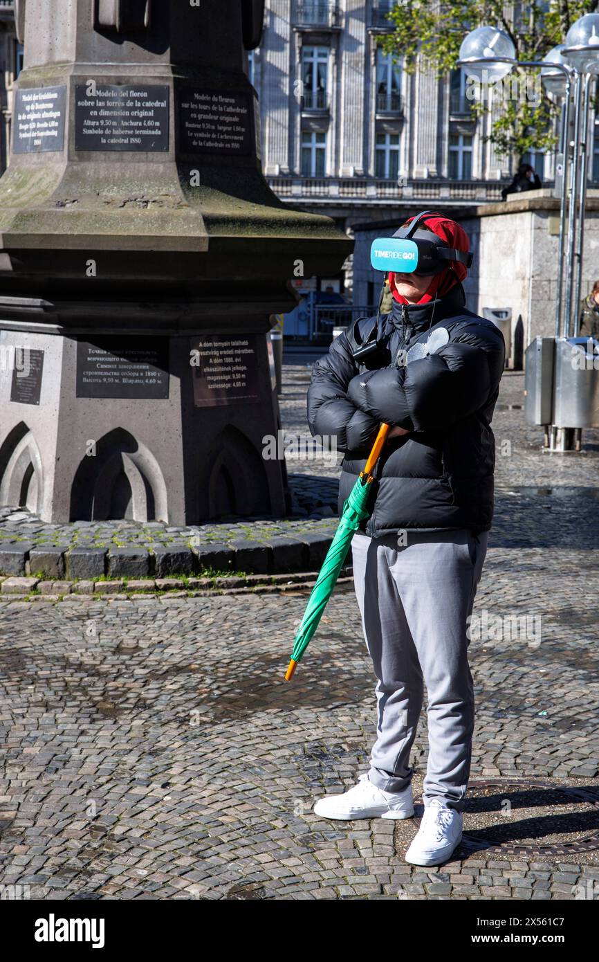 Tourist mit Virtual-Reality-Brille vor dem Kölner Dom Köln, Deutschland. Das Unternehmen TIMERIDE bietet Stadtführungen mit Virtual-Reality-Glas an Stockfoto