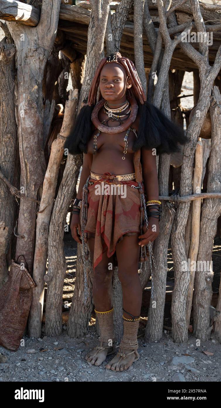 Junge, traditionell gekleidete Himba-Frau, lehnt sich an eine Holzhütte in der Nähe von Opuwo, Kaokoveld, Kunene, Namibia Stockfoto