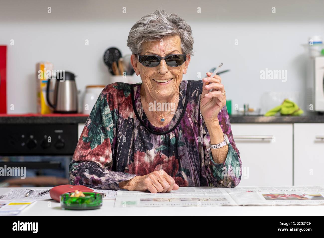 Eine alte Frau mit Falten sitzt in der Küche mit Sonnenbrille und raucht fröhlich eine Zigarette