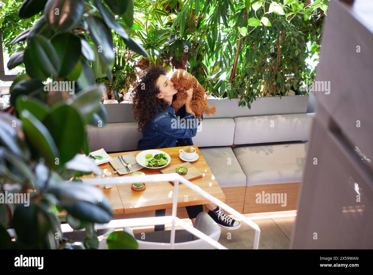 Porträt einer schönen lockigen brünetten Frau, die ihr Haustier umreißt, während sie in einem veganen Restaurant mit umweltfreundlicher Inneneinrichtung ein pflanzliches Essen genießt Stockfoto