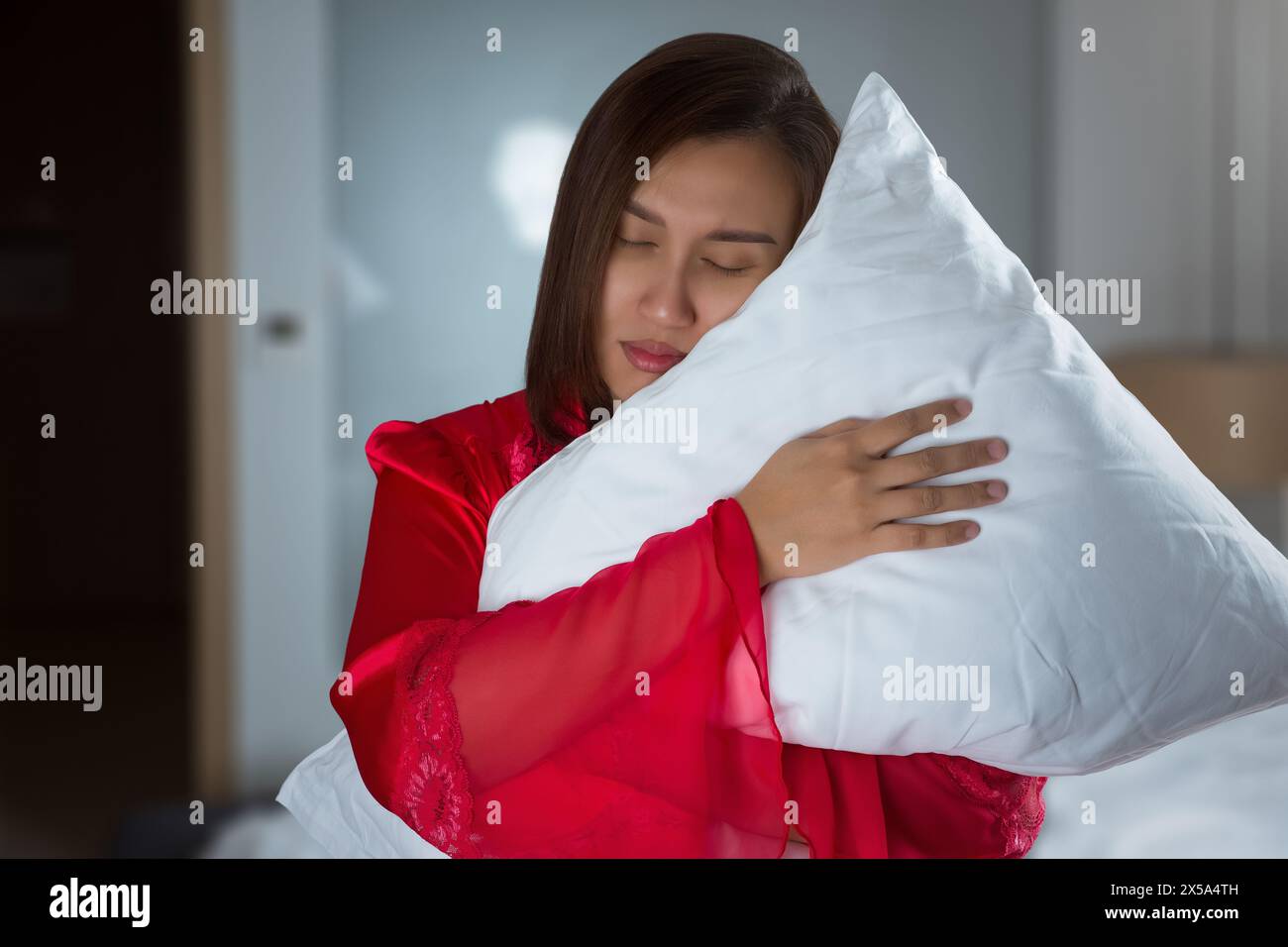 Eine asiatische Frau in einem roten Seidennachthemd schläft beim Stehen ein und umarmt nachts ein Kissen in ihrem Schlafzimmer. Stockfoto
