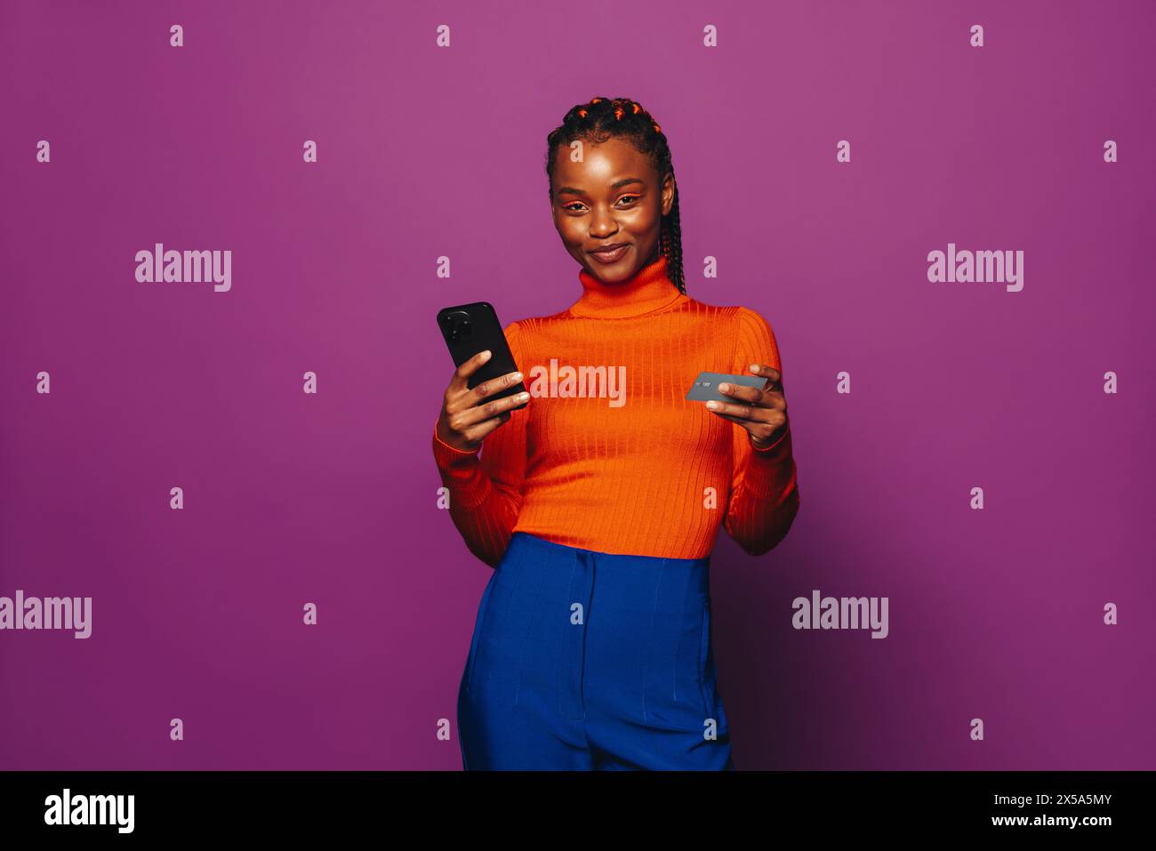 Eine junge Frau mit zweifarbigen Zöpfen steht vor einem violetten Hintergrund und nutzt ein Smartphone, um eine Online-Zahlung über eine Shopping-App zu tätigen. Stockfoto