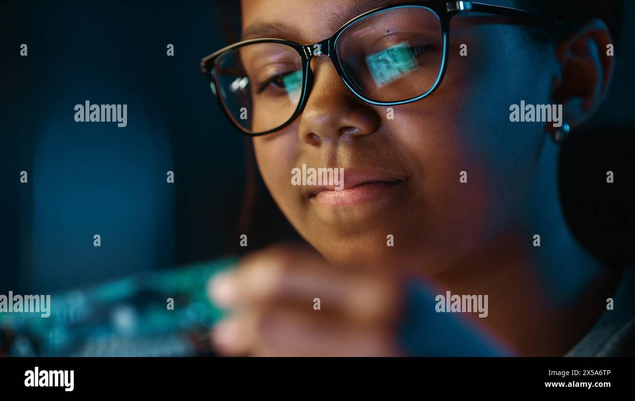 Nahaufnahme Porträt eines süßen jungen, multiethnischen Schwarzen Mädchens im Teenageralter studiert ein kleines Leiterplattenobjekt. Junge Frau trägt Brille und arbeitet an ihrem Schulwissenschaftsprojekt. Stockfoto