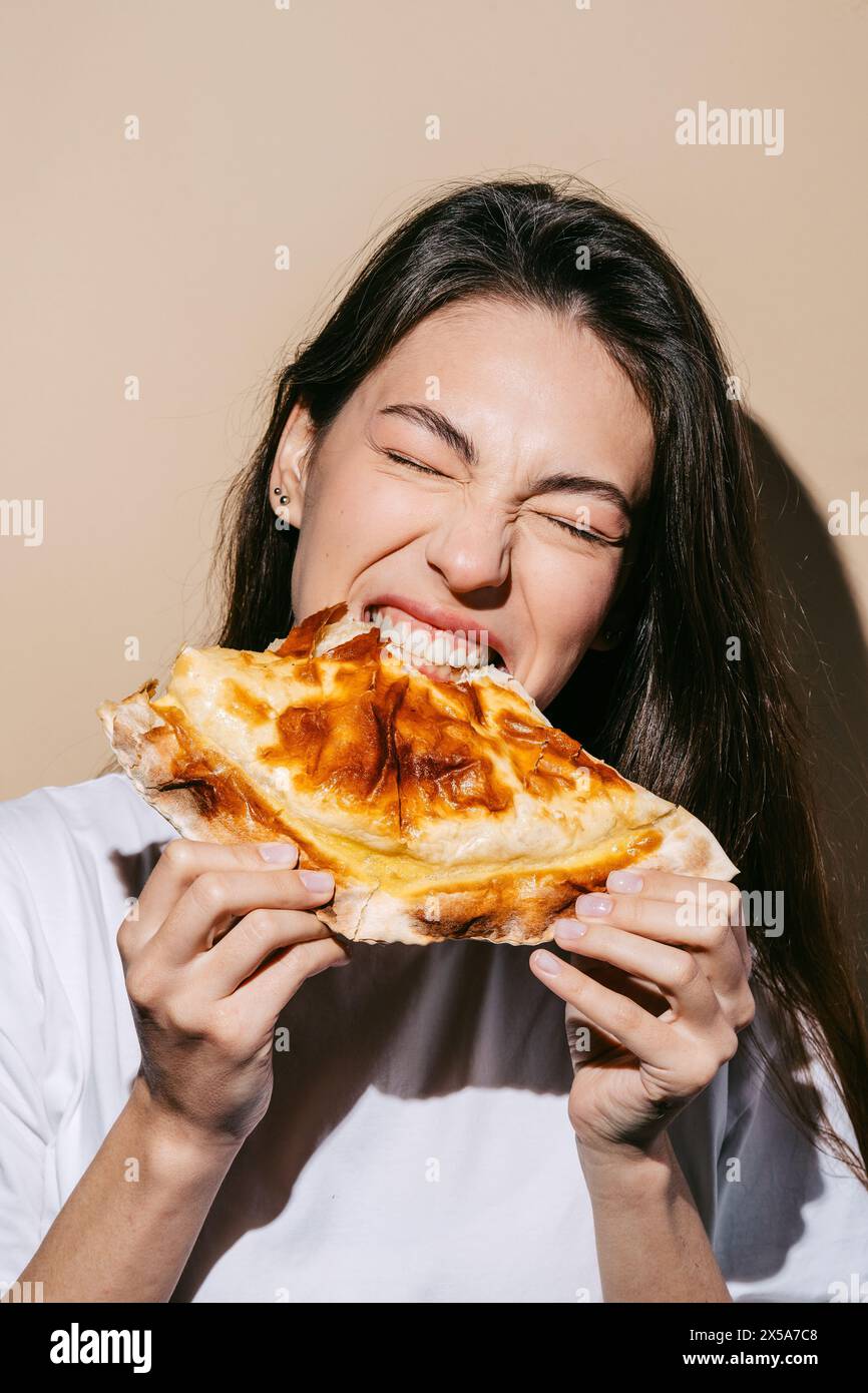 Eine fröhliche Frau beißt in einen frisch gebackenen Cheburek und zeigt die goldbraune Kruste und die herzhafte Füllung. Stockfoto