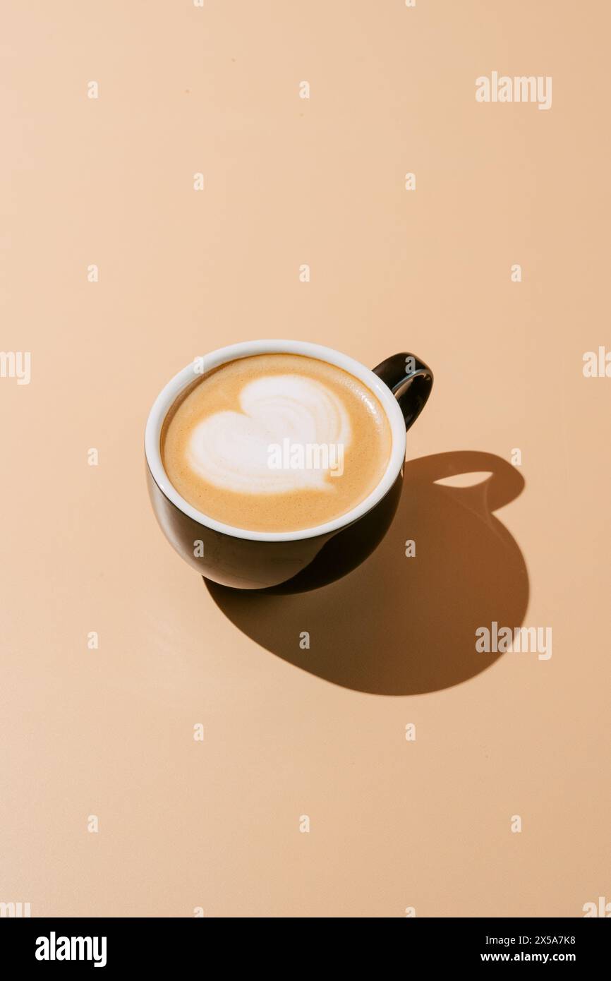 Von oben wunderschön gefertigter Cappuccino mit herzförmiger Latte Art, präsentiert in einem klassischen Keramikbecher mit weichem Schatten auf einem cremefarbenen Hintergrund Stockfoto