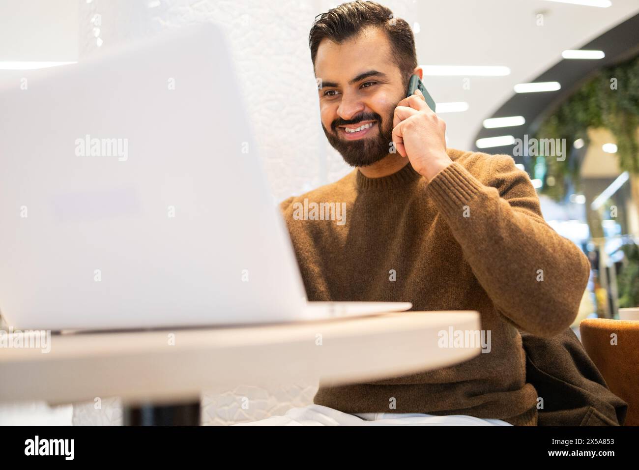 Ein Mann, der engagiert und sympathisch ist, führt ein Telefongespräch mit einem warmen Lächeln, sitzt mit seinem Laptop in einem modernen Café Stockfoto