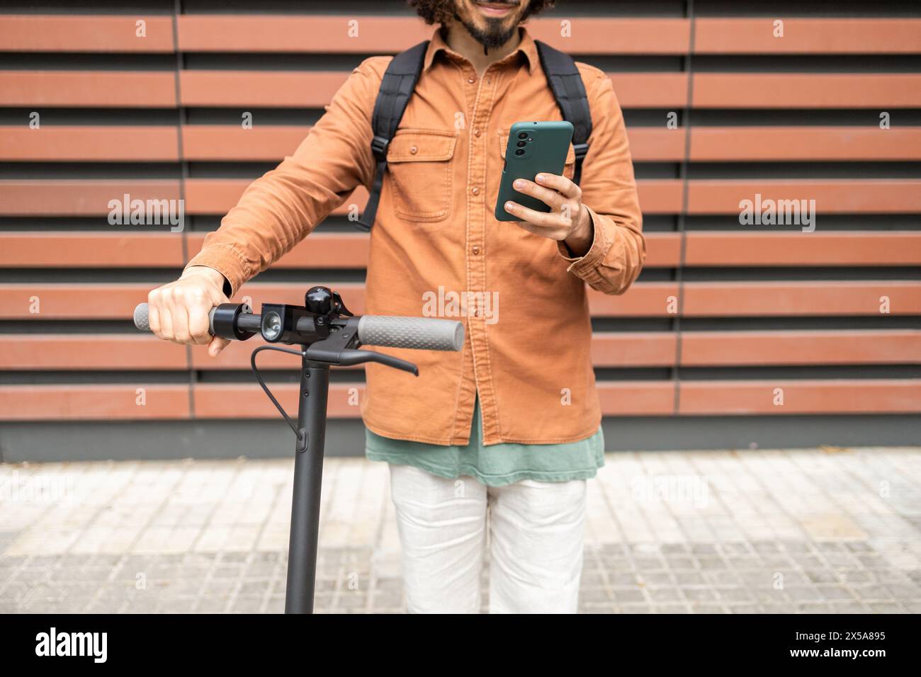 Ein junger Mann in lässiger Kleidung interagiert mit seinem Smartphone, während er sich an einem elektrischen Roller festhält und vor einem geometrischen Hintergrund steht Stockfoto