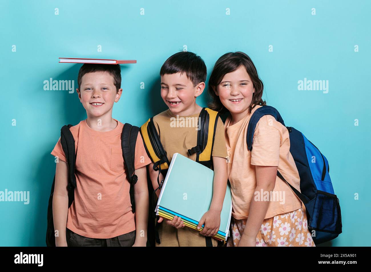 Drei fröhliche Schulkinder, zwei Jungen und ein Mädchen, stehen vor einem türkisfarbenen Hintergrund. Ein Junge balanciert ein Buch auf den Kopf und lächelt den Camer an Stockfoto