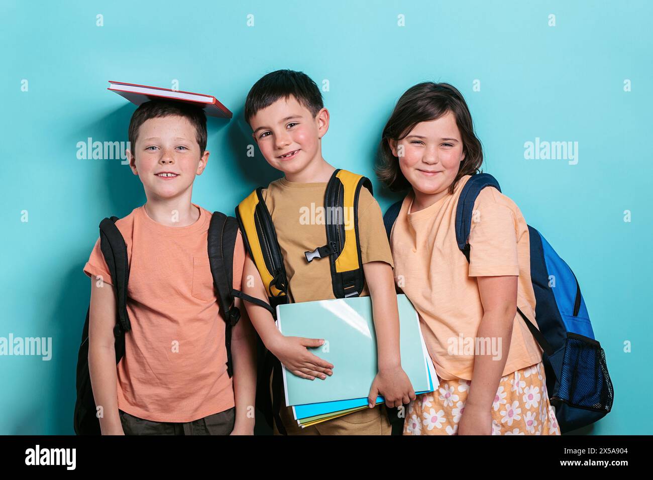 Drei fröhliche Schulkinder, zwei Jungen und ein Mädchen, stehen vor einem türkisfarbenen Hintergrund. Ein Junge balanciert ein Buch auf den Kopf und lächelt den Camer an Stockfoto
