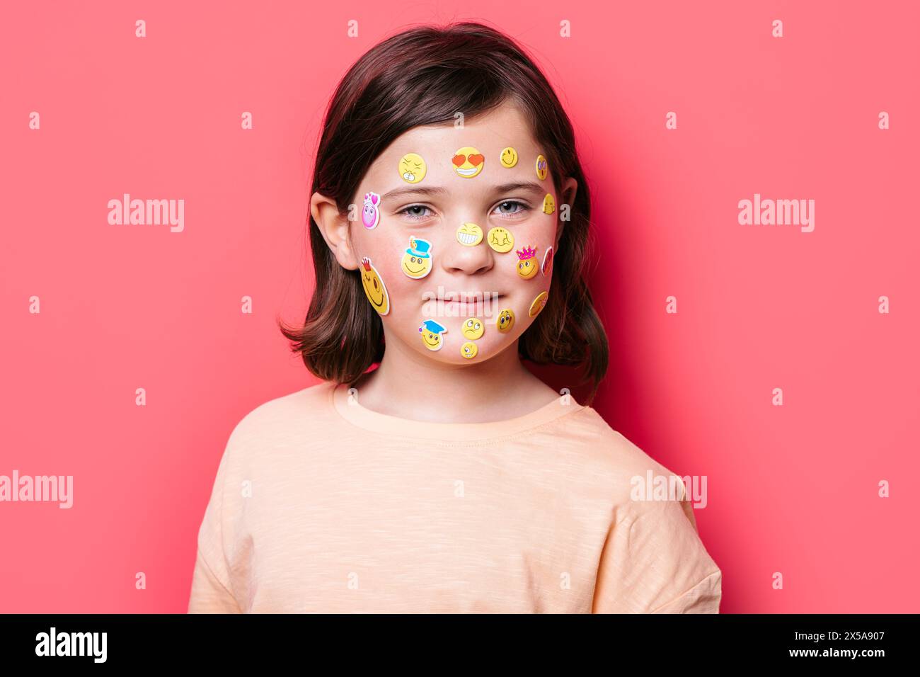 Ein junges Schulmädchen mit einem zufriedenen Lächeln blickt direkt in die Kamera, ihr Gesicht ist verspielt mit bunten Emoji-Stickern an einer leuchtenden Nadel verziert Stockfoto