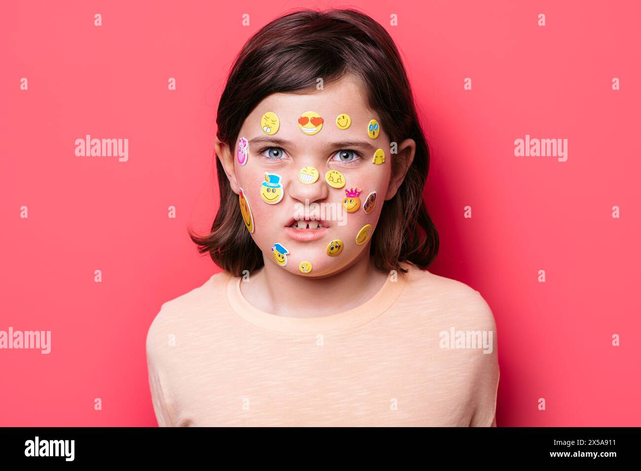 Schulmädchen mit einer Vielzahl von bunten Emoji-Aufklebern auf dem Gesicht, die die Kamera mit einem neutralen Ausdruck vor einem rosafarbenen Hintergrund betrachten. Stockfoto