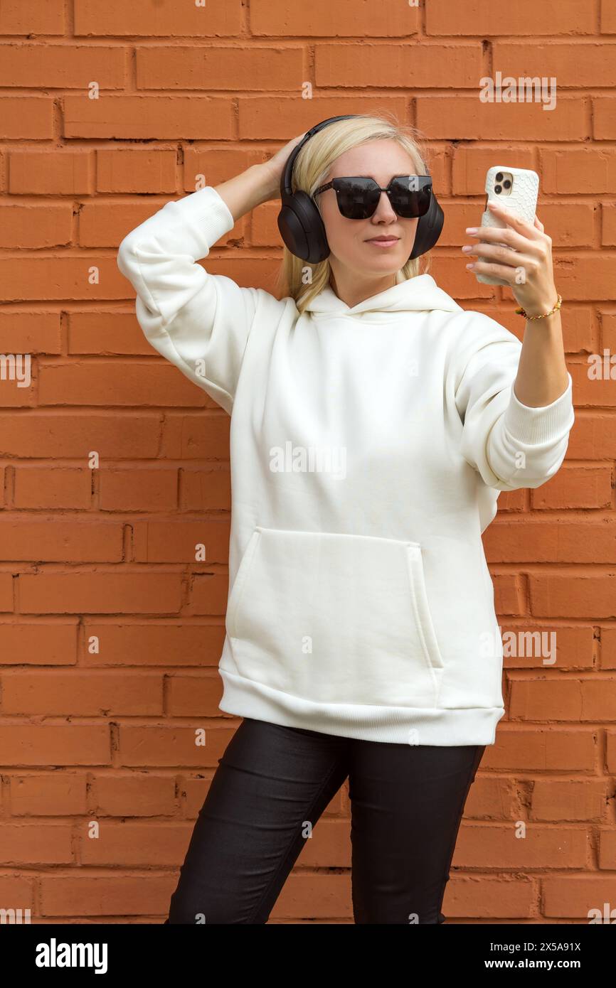 Die blonde Frau in lässiger Kleidung verwendet ihr Smartphone, während sie Kopfhörer gegen eine städtische Ziegelwand trägt. Stockfoto