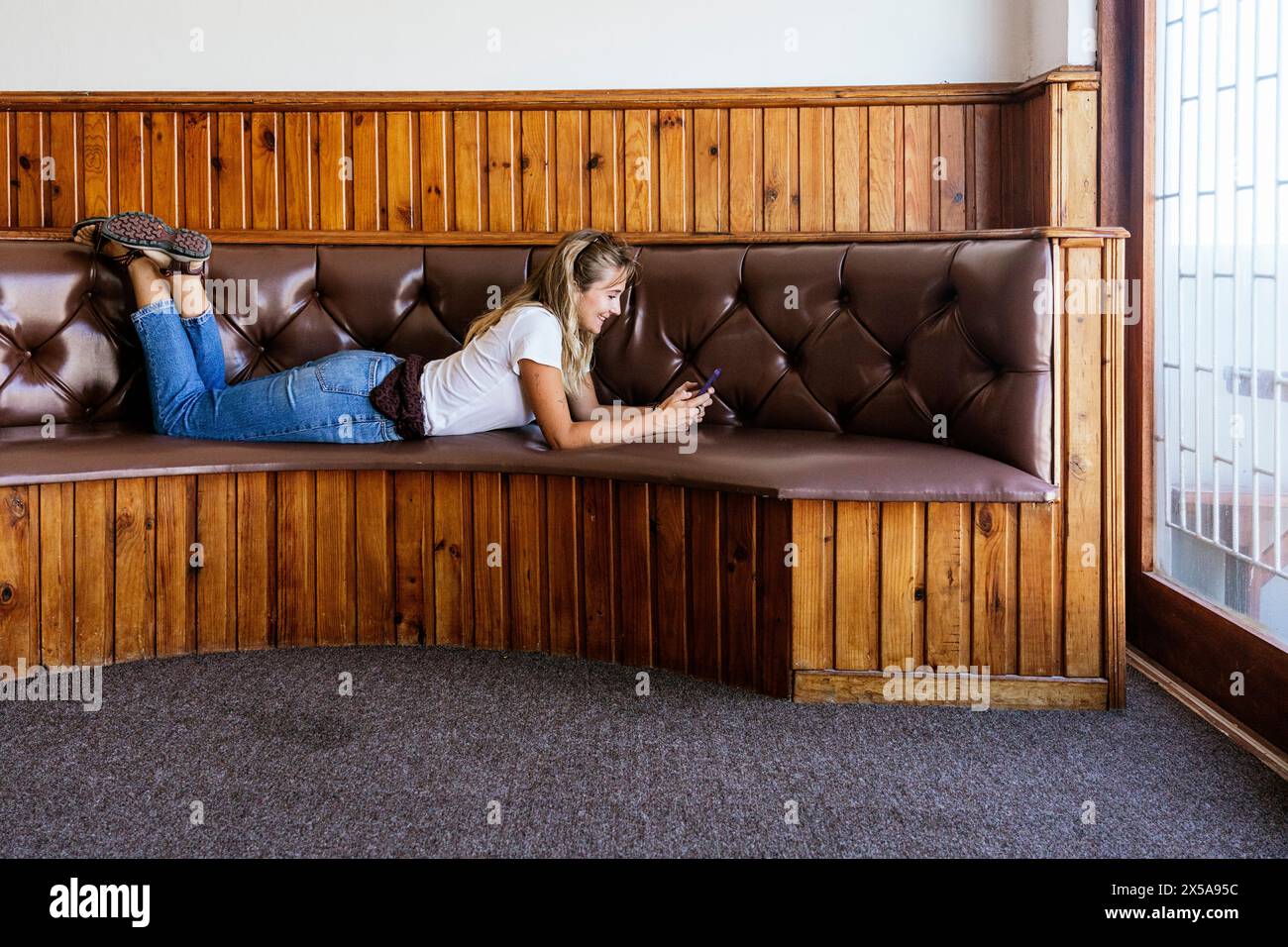 Junge Frau, die sich entspannt und ihr Smartphone auf einer Vintage-Lederbank mit Holzvertäfelung benutzt. Stockfoto