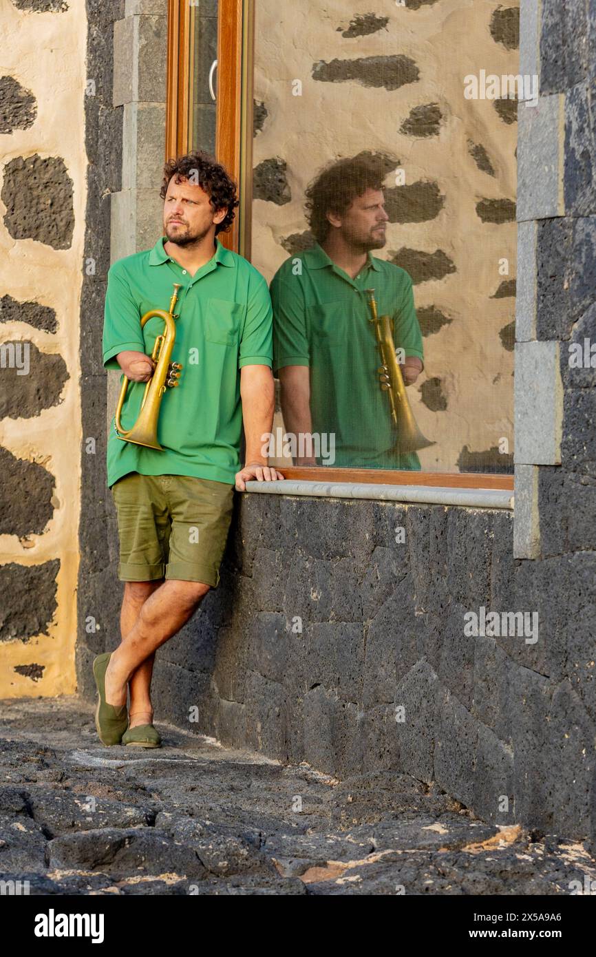 Ein einarmiger Mann mit einer Trompete steht neben einem reflektierenden Fenster und blickt weg, zeigt Widerstandskraft und musikalische Leidenschaft. Stockfoto