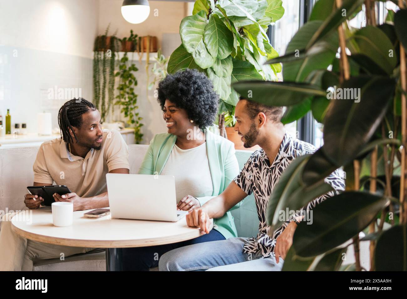 Drei Profis arbeiten an einem Laptop in einer modernen, anlagenreichen Coworking-Umgebung zusammen, tauschen Ideen aus und genießen ein freundliches Gespräch Stockfoto