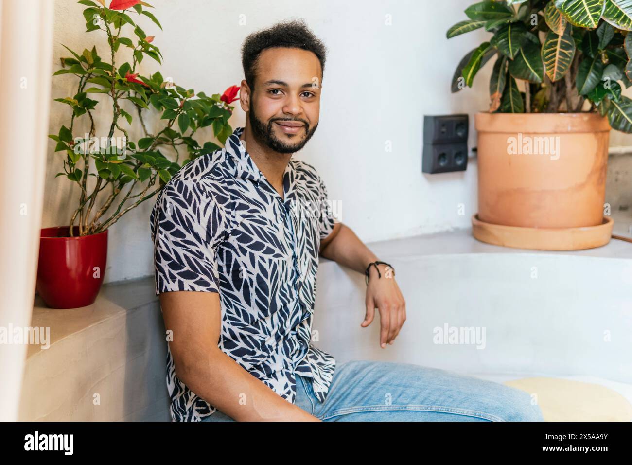 Ein junger Mann mit einem einladenden Lächeln, der lässig in einer kooperativen Umgebung sitzt, umgeben von üppigem Grün Stockfoto
