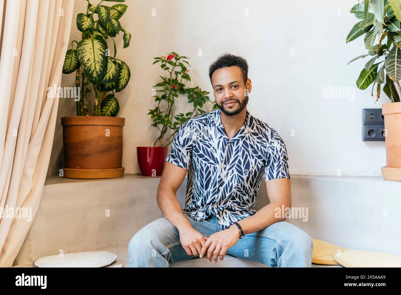 Ein fröhlicher junger Mann saß lässig inmitten von Zimmerpflanzen und verkörperte ein entspanntes Coworking-Umfeld Stockfoto