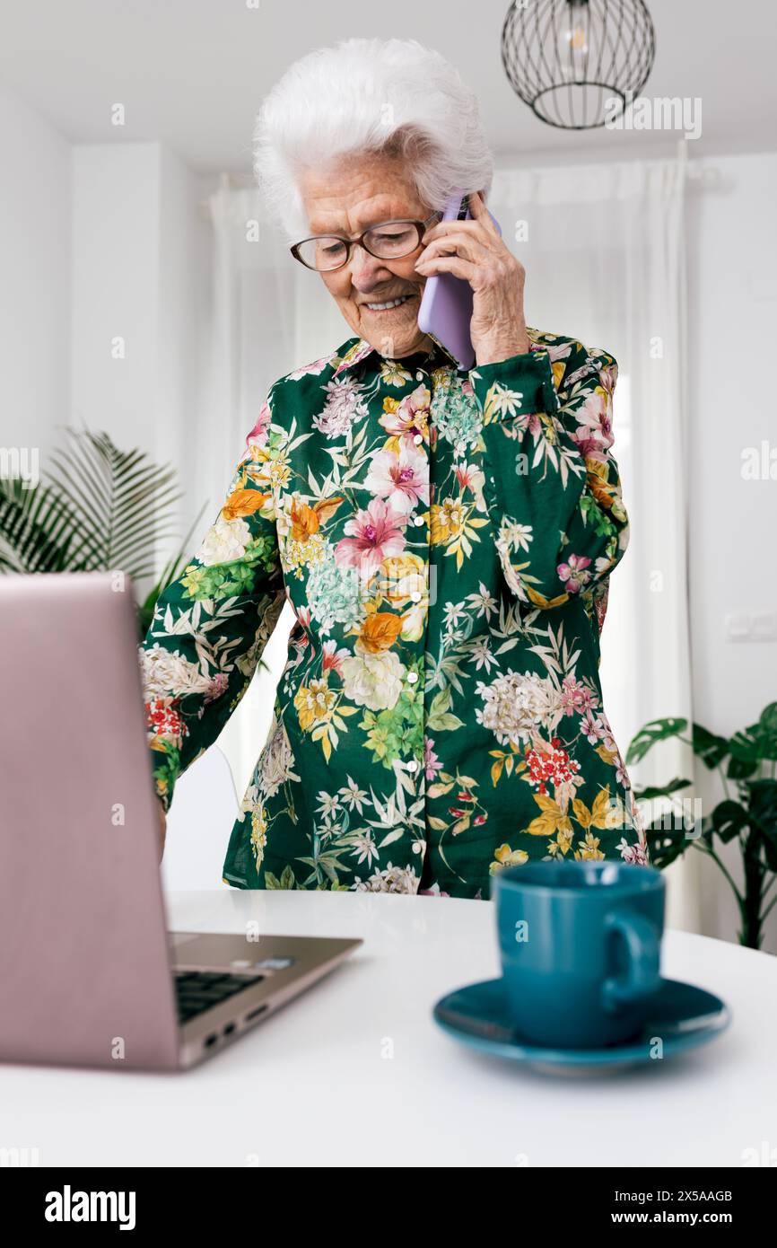 Eine ältere Frau in einer Blumenbluse arbeitet von zu Hause aus, benutzt einen Laptop und unterhält sich am Telefon Stockfoto