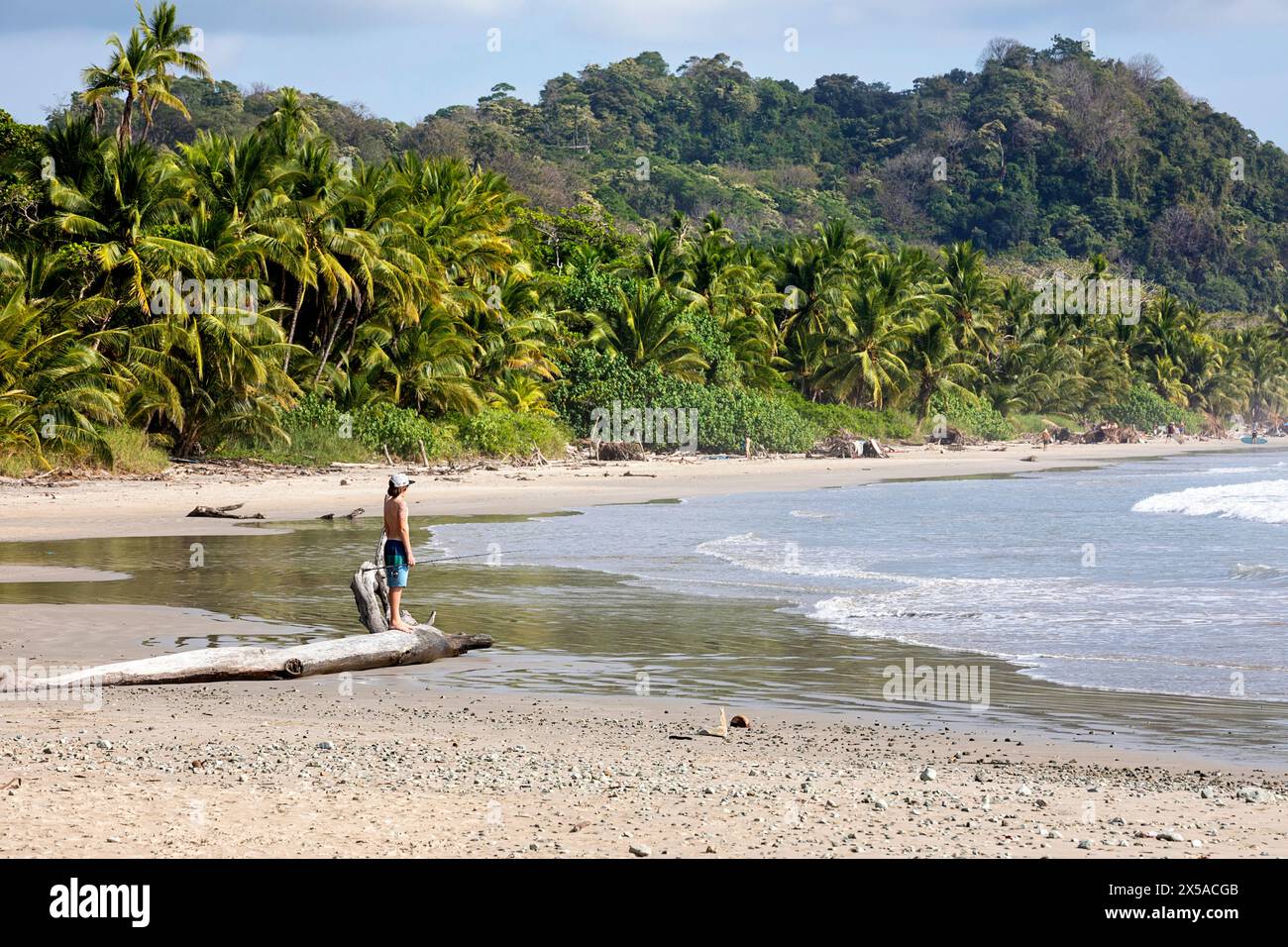 Kind, Tourist mit Angelrute auf einem Baumstamm auf einer Playa Hermosa, Strand auf Nicoya Halbinsel, mit Palmen und tropischem Wald auf der Rückseite, Costa Rica Stockfoto