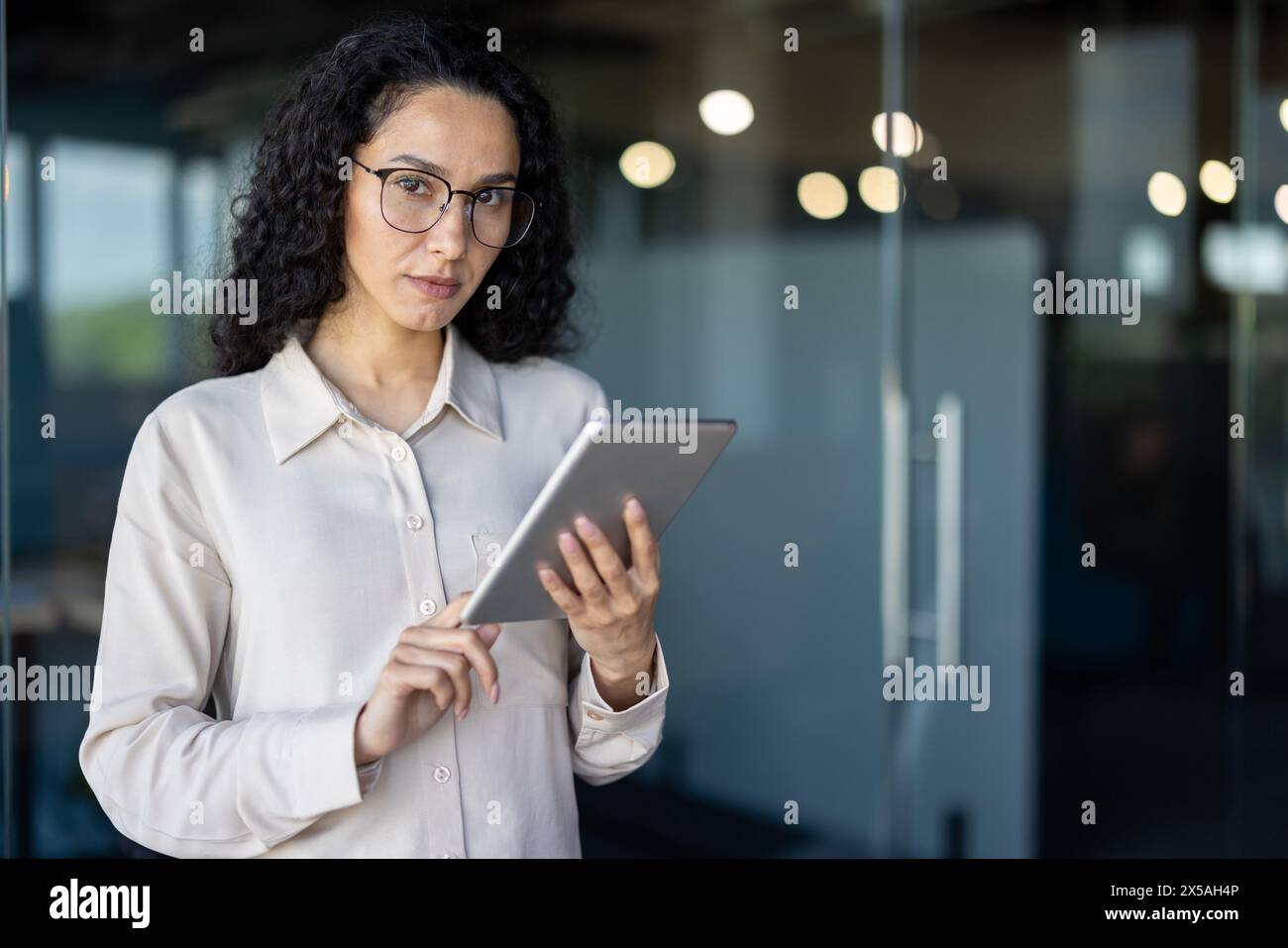 Eine hispanische Geschäftsfrau in einer hellen Bluse verwendet in einer Büroumgebung ein digitales Tablet, um möglicherweise Daten zu überprüfen oder Aufgaben zu verwalten. Stockfoto