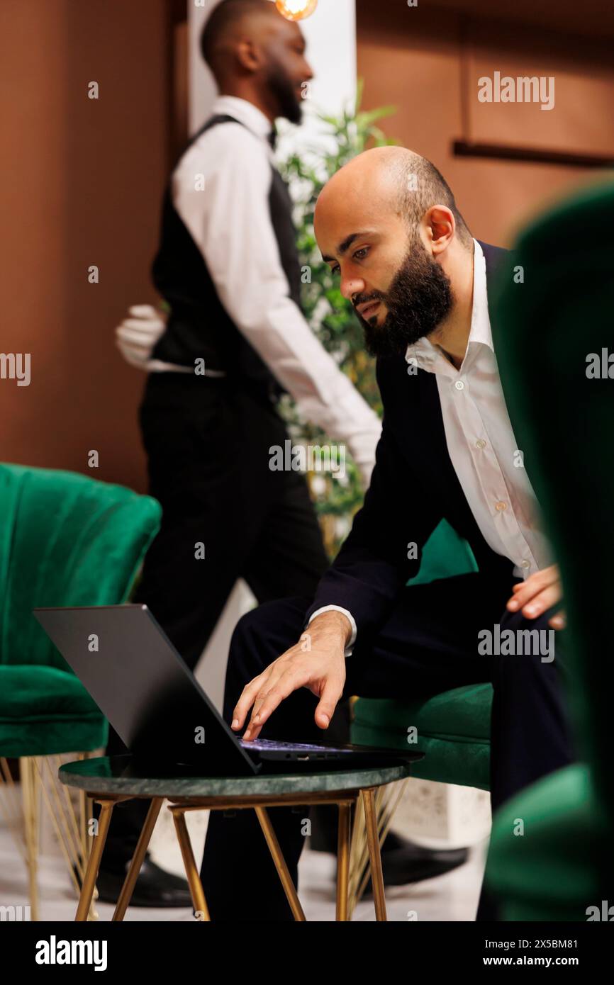 Nahöstlicher Geschäftsmann in Anzug, der sich auf die offizielle Konferenz mit dem Vorstand vorbereitet, Geschäftsreise. Junger Unternehmer, der im Loungebereich an einem Laptop arbeitet, Management-Meeting. Stockfoto