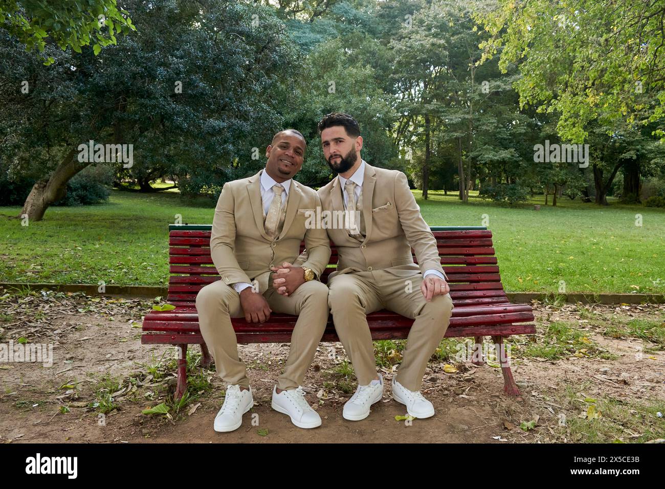 General Shoot von zwei frisch verheirateten Männern, die in die Kamera lächeln, sitzen auf einer Holzbank in einem Garten. Das Konzept von Glück, Schwulenstolz Stockfoto