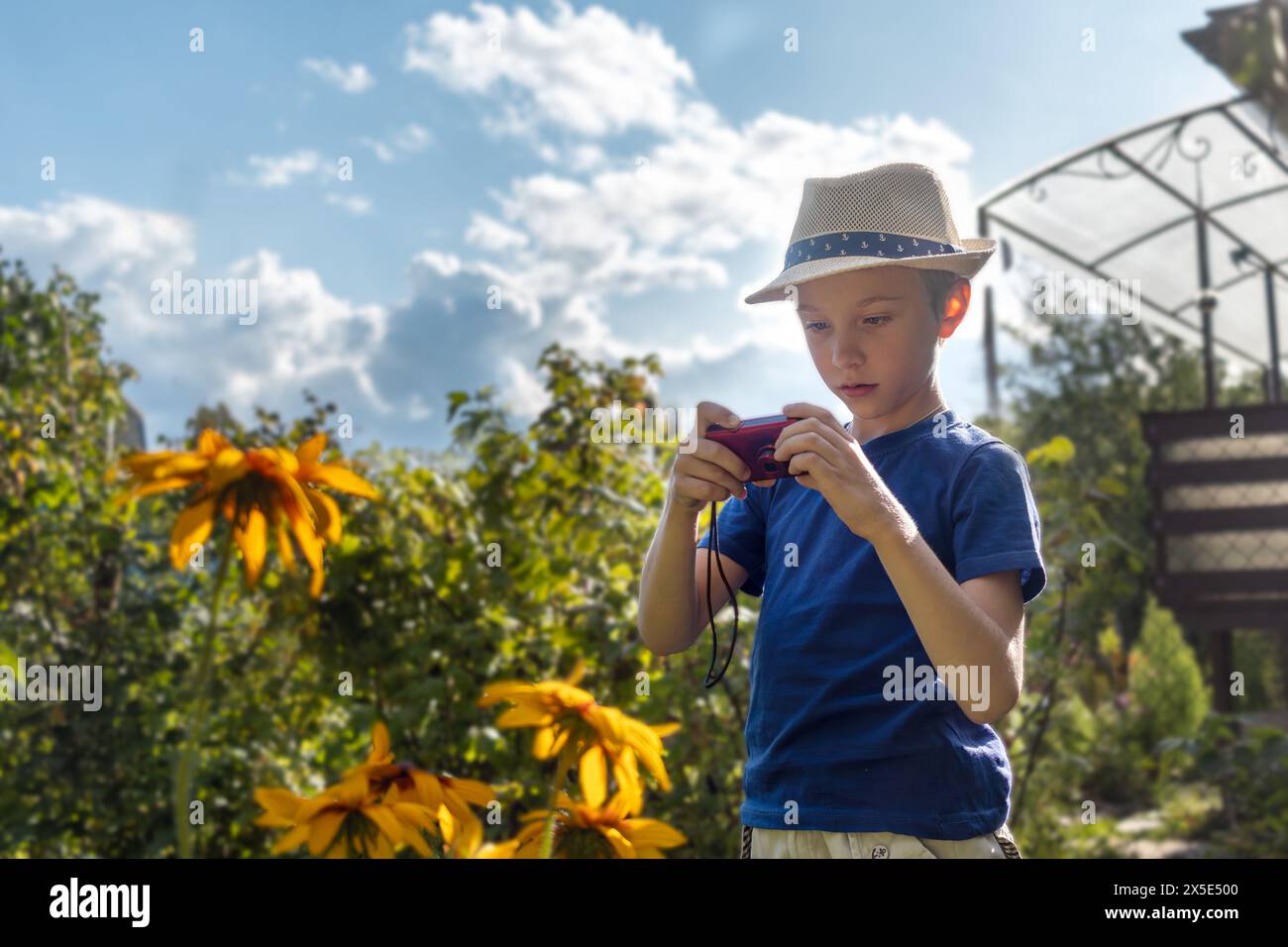 Junge mit Strohhut fotografiert Blumen im Landgarten am sonnigen Sommernachmittag. Interessanter Urlaub, Hobby, Fotografie. Stockfoto