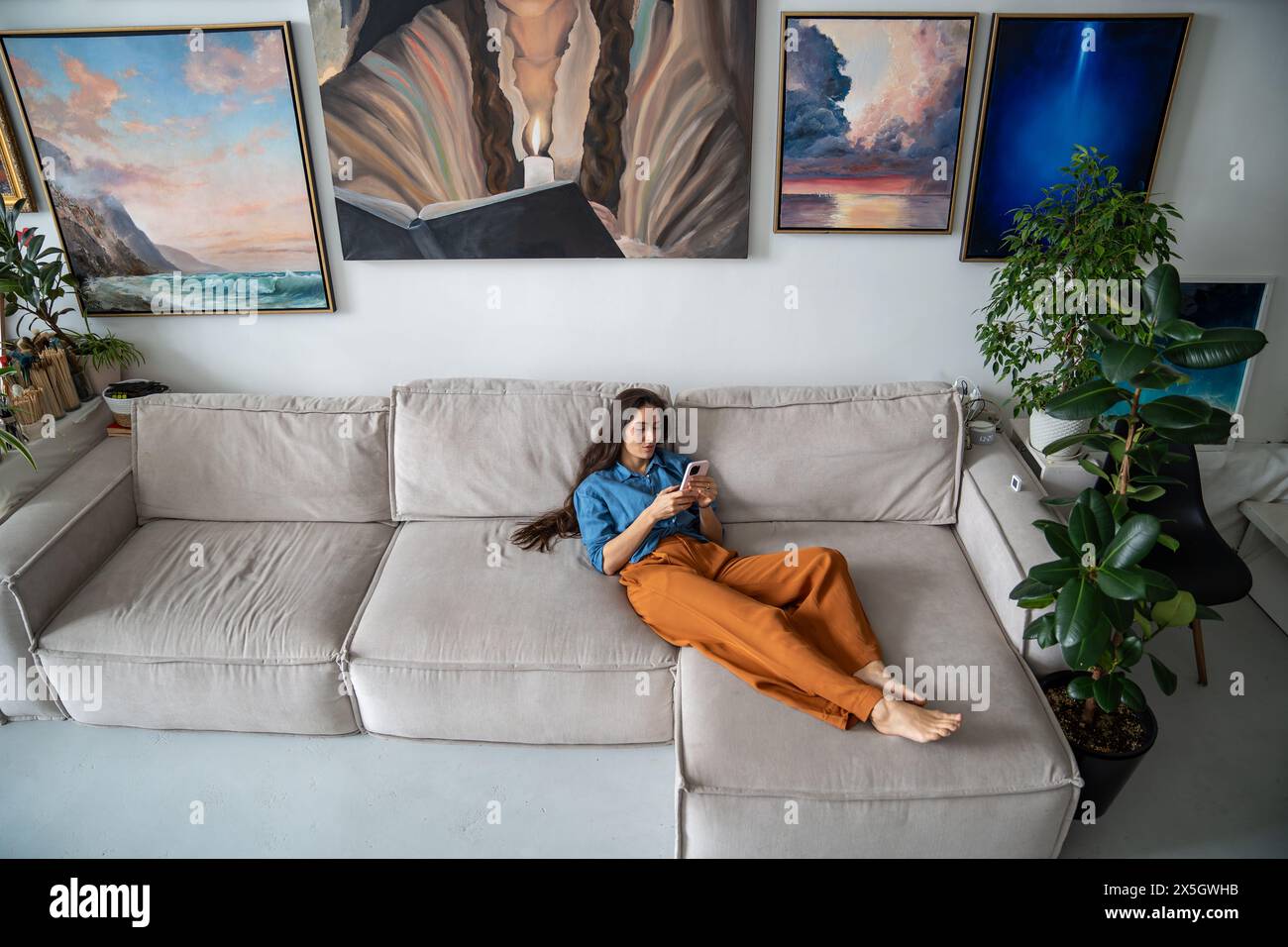 Schlampiges Mädchen, das mit Smartphone auf dem Sofa liegt. Frau, die sich im Kunststudio zu Hause entspannt. Stockfoto