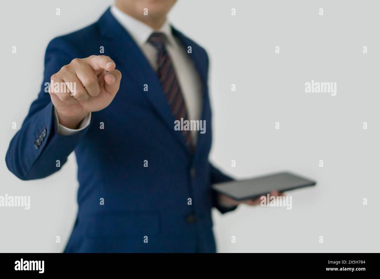 Ein Geschäftsmann in elegantem blauen Anzug und Krawatte, der ein Tablet hält, auf die Kamera zeigt und den Eindruck erweckt, als würde er einen präsentieren oder betonen Stockfoto