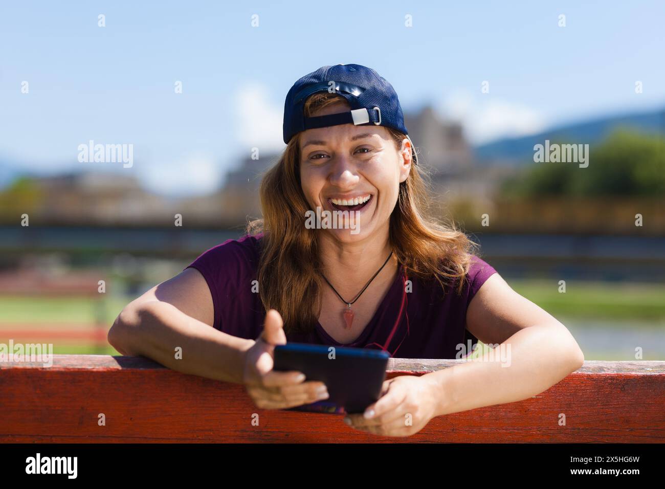 Eine fröhliche junge Frau mit Baseballmütze sitzt draußen und lacht, während sie an einem sonnigen Tag in der Nähe eines Flusses ihr Smartphone ansieht. Stockfoto