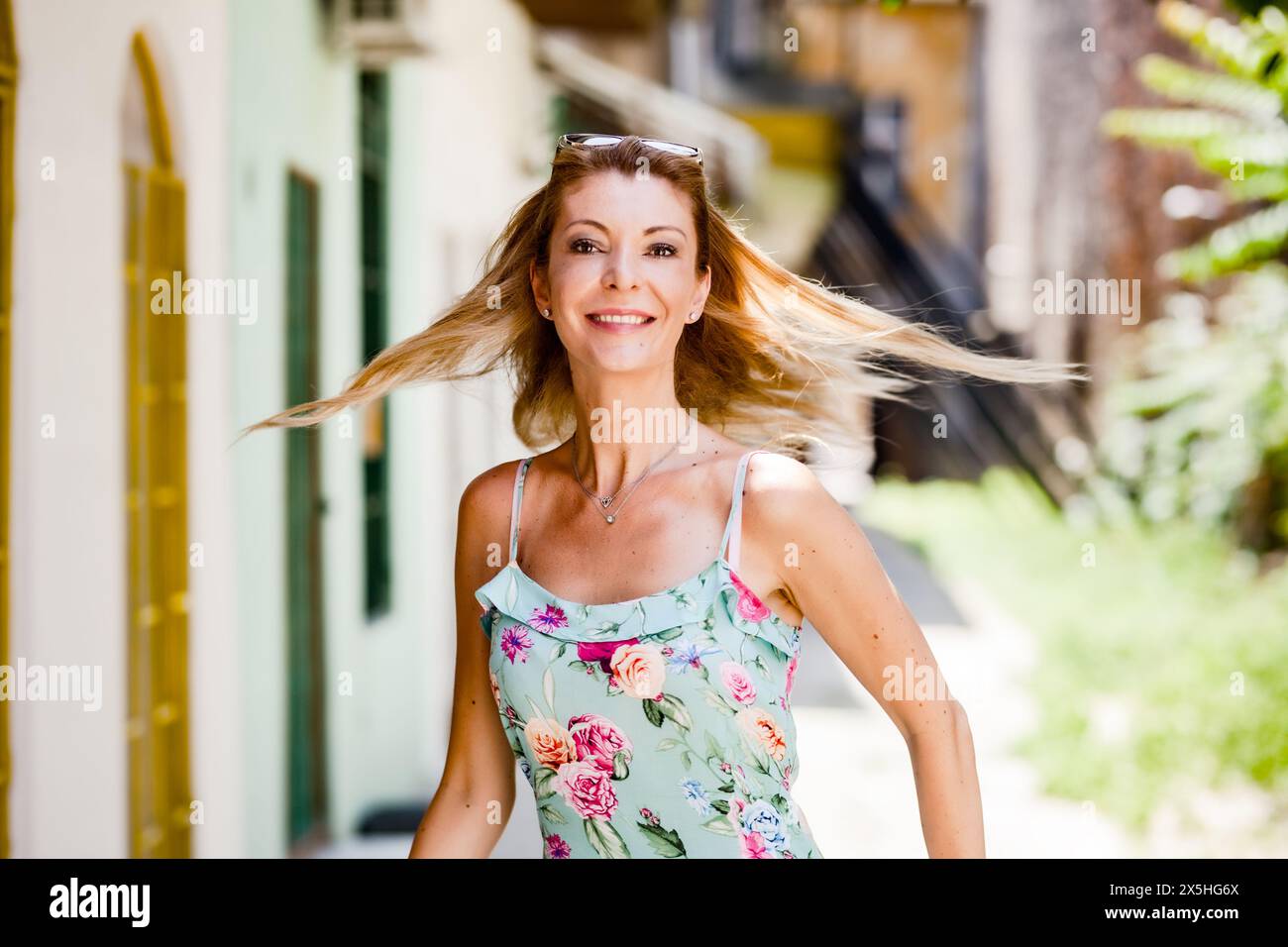Eine fröhliche Frau mittleren Alters mit fließenden Haaren tritt selbstbewusst auf einer sonnendurchfluteten Straße entlang, gekleidet in einem leuchtenden Tanktop mit Blumenmuster. Stockfoto