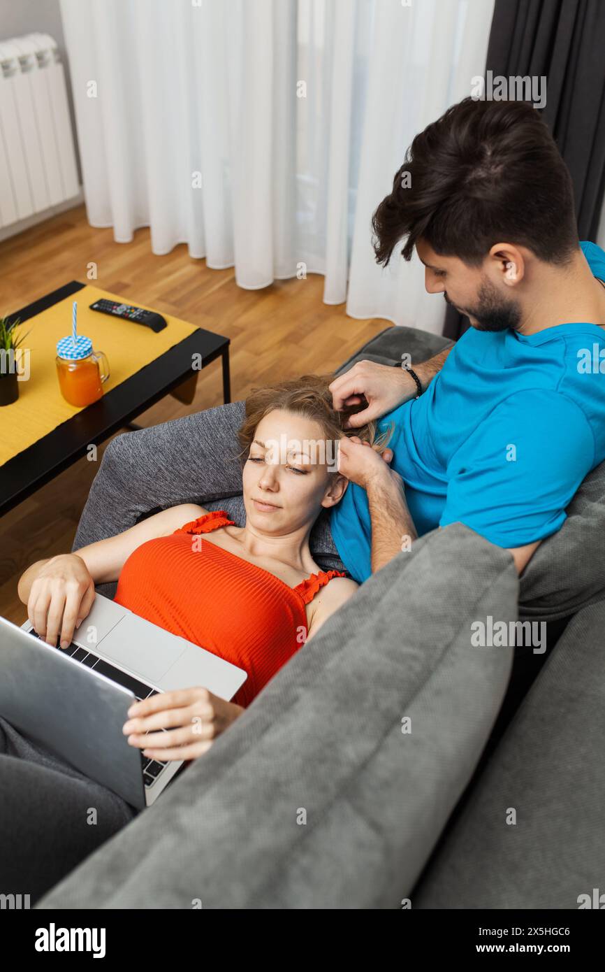 Entspannte und ungezwungene Atmosphäre, während ein junger Mann liebevoll über die Haare seiner Partnerin streichelt, während sie auf einem Laptop sitzt und bequem auf einem Sofa sitzt. Stockfoto