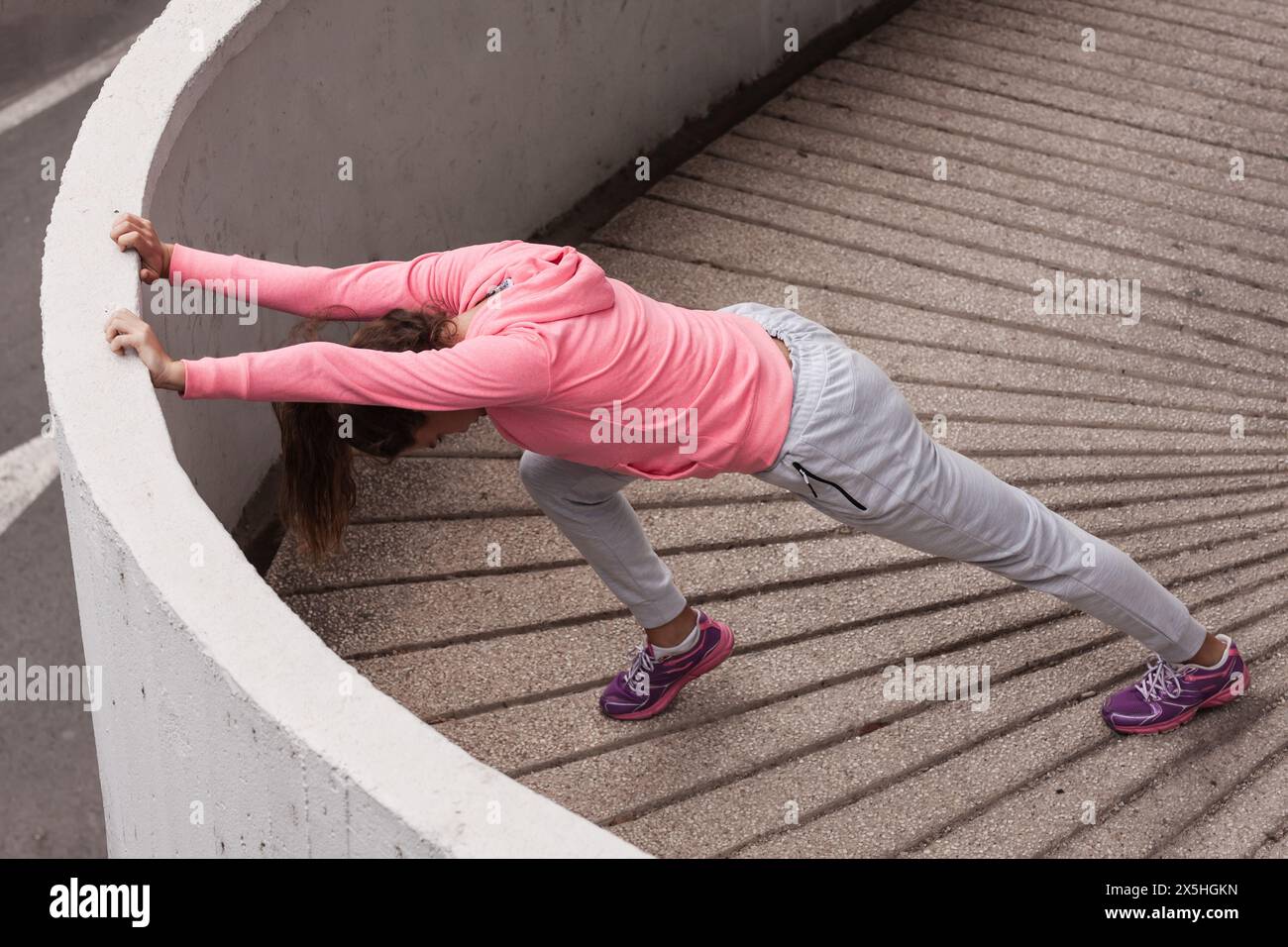 Fokussierte, sportliche Frau in rosa Oberteil und grauer Hose, die Stretchanlagen auf Treppen im Freien ausführt, demonstriert Flexibilität und Fitness-Routine. Stockfoto