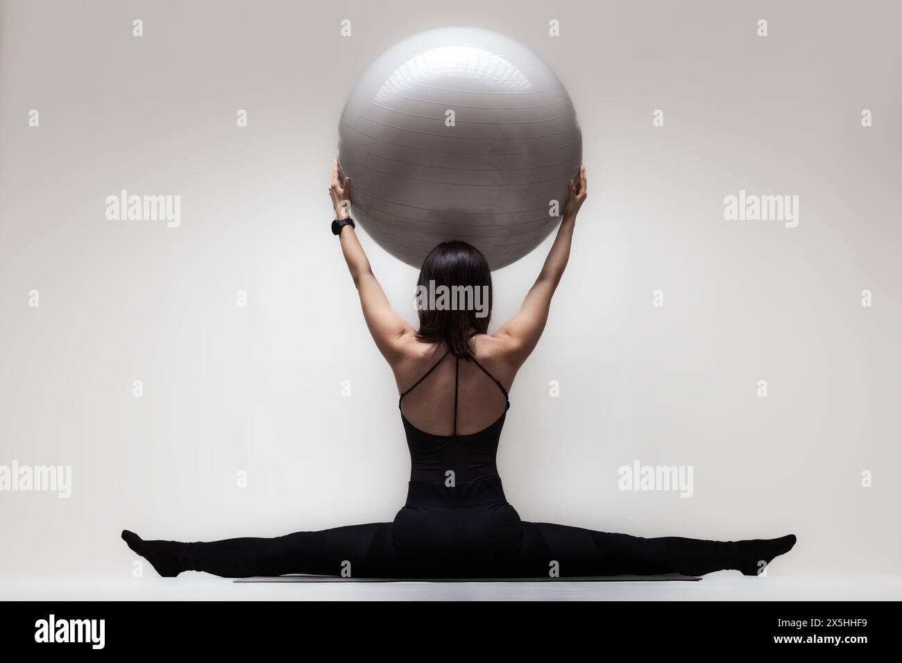 Eine junge Frau in gespaltener Position mit einem großen Fitnessball für Balance und Flexibilität vor neutralem Hintergrund. Stockfoto