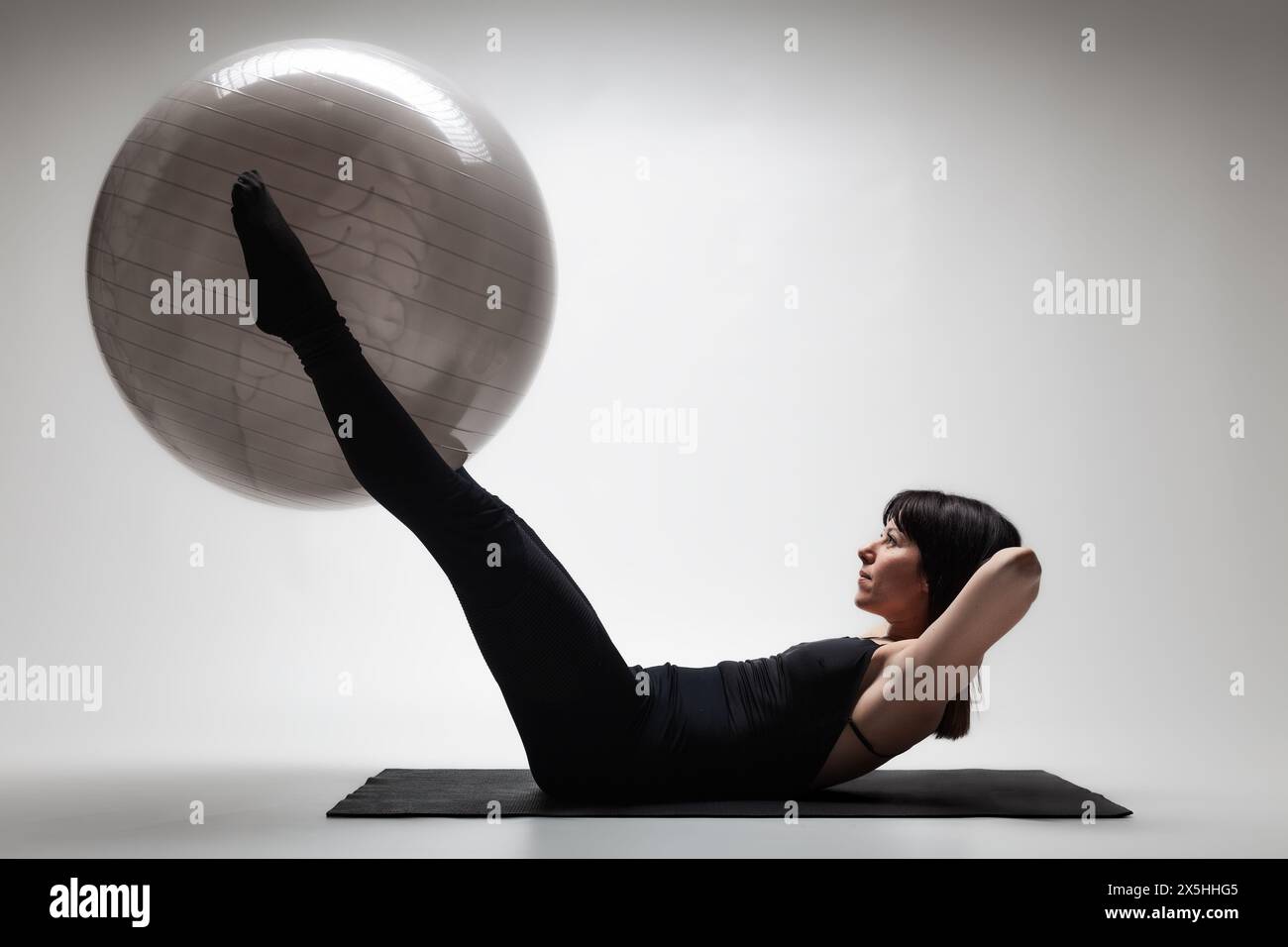 Eine fokussierte junge Frau nimmt an einer Pilates-Sitzung Teil, indem sie einen großen Fitnessball auf einer Yoga-Matte in einer sanft beleuchteten Studioumgebung verwendet. Stockfoto