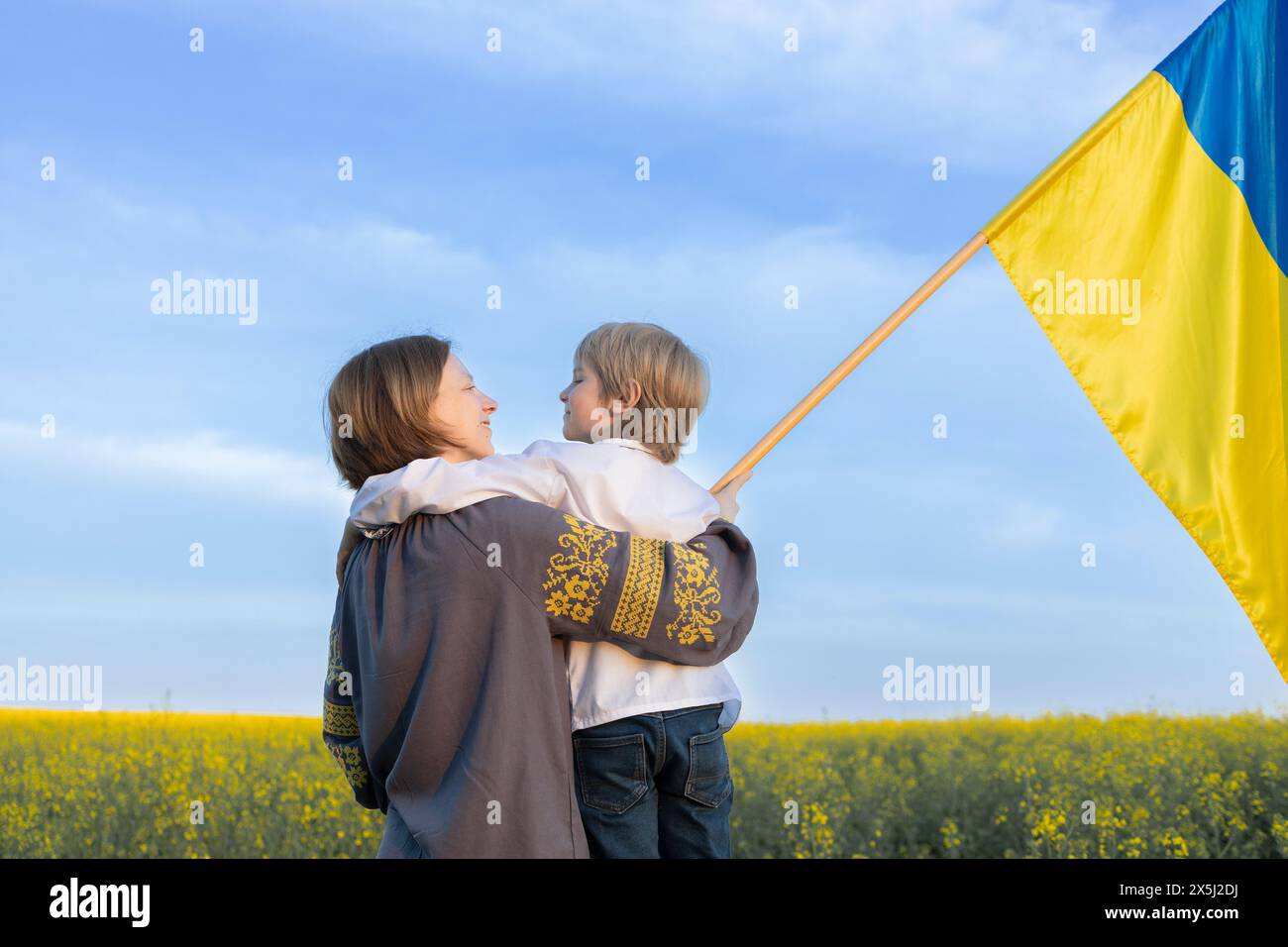 Ukrainische Familie, Mutter und Sohn mit blauer und gelber Flagge inmitten eines blühenden Rapsfeldes. Unabhängigkeitstag der Ukraine. Unterstützung der Ukraine bei der Konfrontation Stockfoto