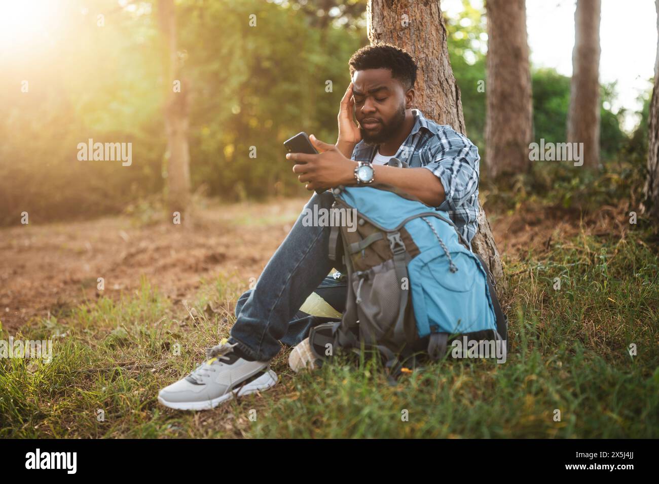 Ein besorgter Wanderer hat sich verirrt und er benutzt ein Telefon, um die richtige Richtung zu finden, während er in der Natur sitzt. Stockfoto