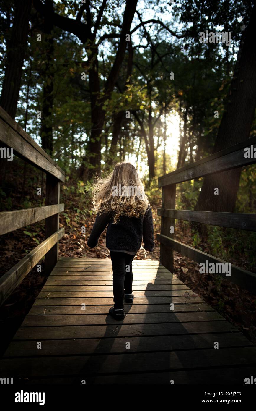 Ein junges Mädchen mit blonden Locken, das im grünen Wald einen Holzweg entlang geht Stockfoto