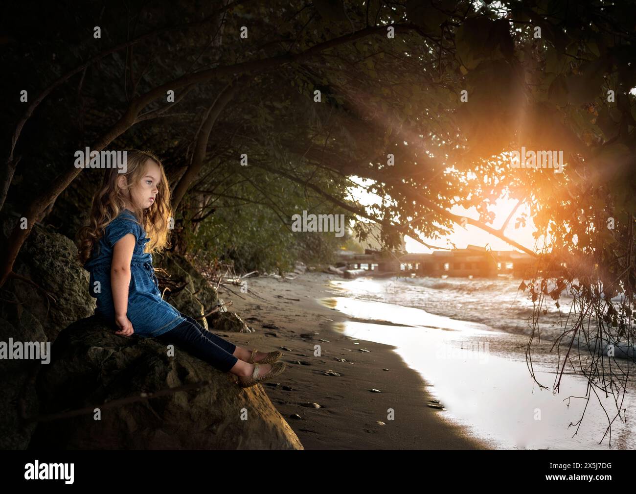 Kleines Mädchen mit langen blonden Locken, das unter sonnigem Himmel auf das Wasser blickt Stockfoto