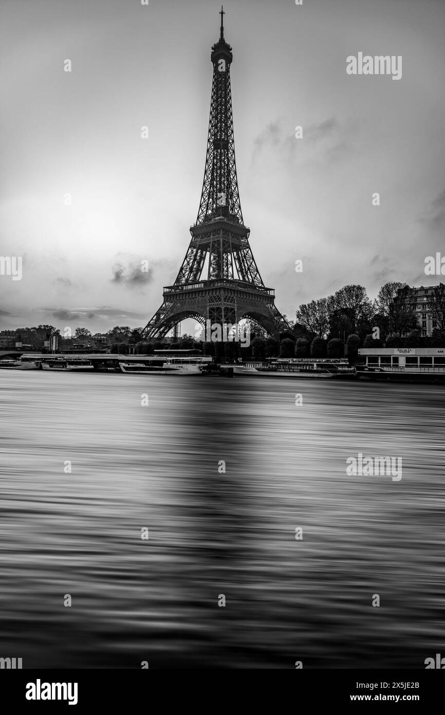 Ein fesselndes Schwarz-weiß-Bild, das den legendären Eiffelturm vor einem bewölkten Himmel zeigt, während die seine im Vordergrund sanft fließt und Paris zeitlose Schönheit zeigt. Frankreich. Schwarzweißbild. Stockfoto