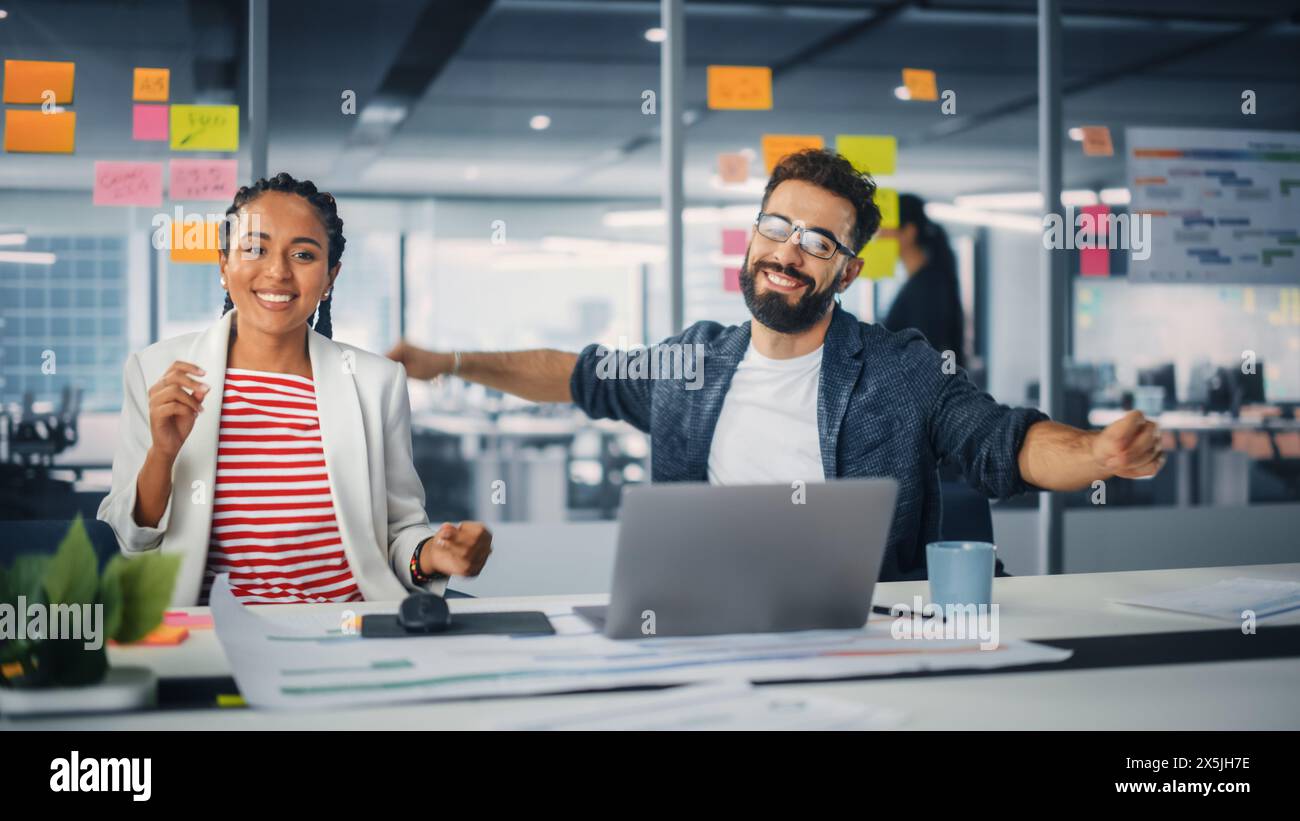 Zwei Happy Young Professionals im Büro: Schwarze Geschäftsfrau und hispanische Geschäftsmann tanzen zusammen, lächeln und in die Kamera schauen, am Schreibtisch sitzen. Spezialisten arbeiten in Creative Office, feiern Erfolge Stockfoto