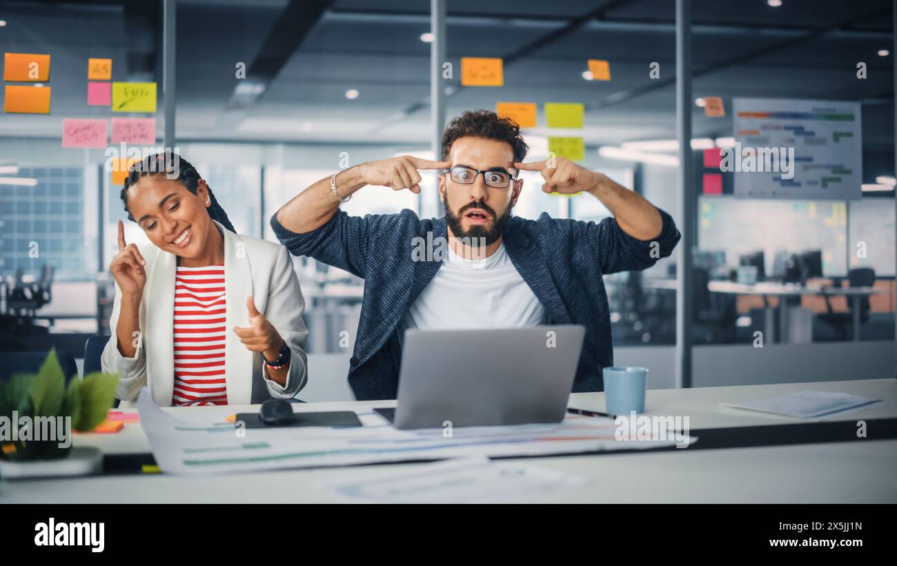 Zwei Happy Young Professionals im Büro: Schwarze Geschäftsfrau und hispanische Geschäftsmann tanzen zusammen, lächeln und in die Kamera schauen, am Schreibtisch sitzen. Spezialisten arbeiten in Creative Office, feiern Erfolge Stockfoto